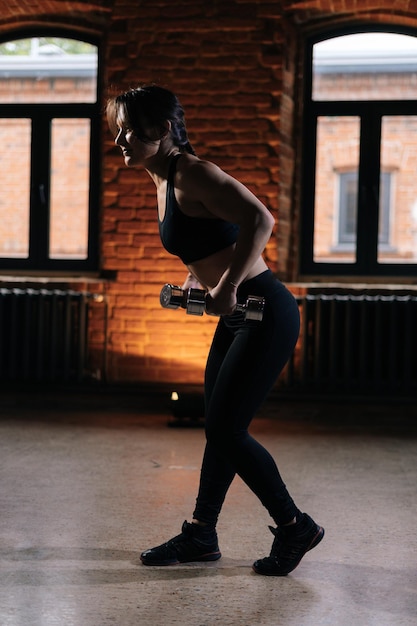 Vista lateral de una joven atlética musculosa con un hermoso cuerpo fuerte usando ropa deportiva trabajando con pesas. Entrenamiento femenino de fitness caucásico haciendo ejercicio en el gimnasio oscuro.