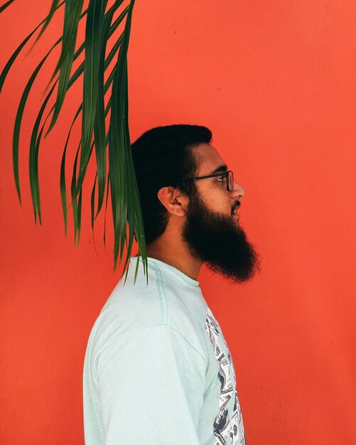 Foto vista lateral de un joven con barba de pie contra una pared naranja