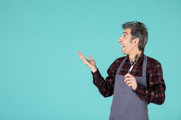 Vista lateral del joven barbero preguntándose con delantal gris y sosteniendo el peine en la cabeza y mostrando la tijera preguntando sobre algo sobre fondo de color azul