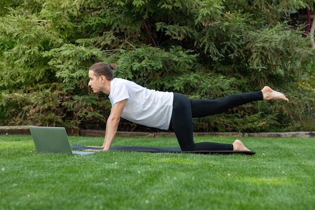 Vista lateral del joven en un bollo en el jardín usando una computadora portátil para aprender a hacer yoga en una clase magistral en línea