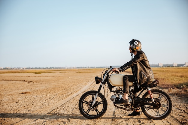 Vista lateral de un joven brutal con una chaqueta negra y gafas conduciendo una motocicleta retro