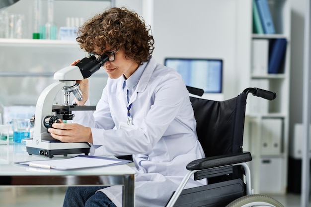 Vista lateral del joven científico en bata de laboratorio sentado en silla de ruedas por lugar de trabajo