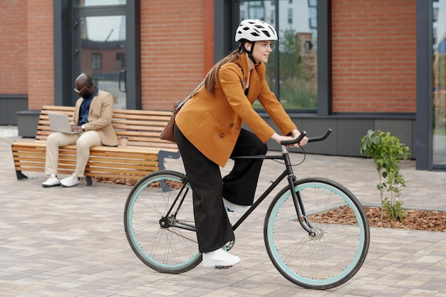 Vista lateral de una joven y elegante empresaria sentada en bicicleta y yendo al trabajo