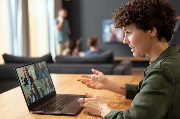 Foto vista lateral de la joven empresaria contemporánea sentada junto a la mesa frente a la computadora portátil y comunicándose con sus colegas en el chat de video en casa