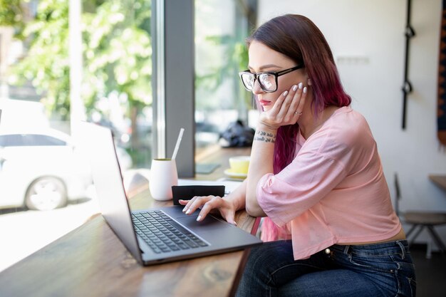 Vista lateral de una joven empresaria sentada a la mesa en una cafetería en la mesa con una taza de café y una computadora portátil