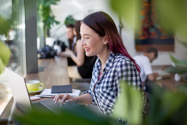 Vista lateral de la joven empresaria sentado a la mesa en la cafetería en la mesa taza de café y portátil i ...