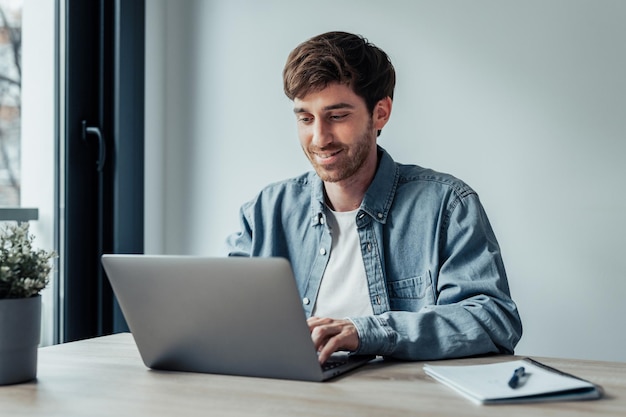 Vista lateral joven empresario guapo que trabaja con computadora remotamente sentado en una mesa de madera en la oficina agradable hombre feliz comunicándose en la red social buscando información en línea xA