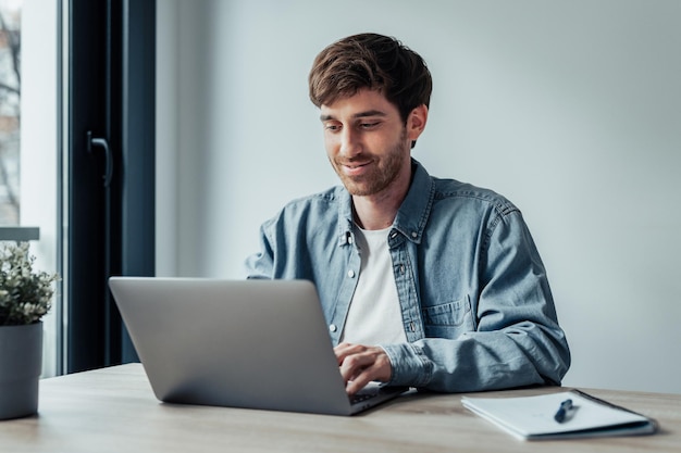 Vista lateral joven empresario guapo que trabaja con computadora remotamente sentado en una mesa de madera en la oficina agradable hombre feliz comunicándose en la red social buscando información en línea xA