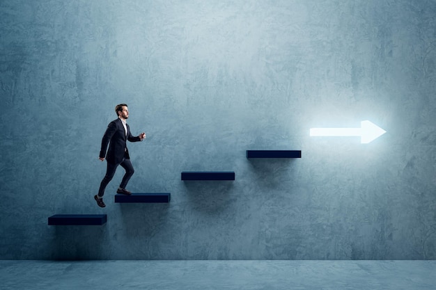 Vista lateral de un joven empresario que sube las escaleras de flecha iluminadas hacia el éxito en el fondo de la pared de hormigón en el interior Desarrollo profesional de crecimiento financiero y concepto avanzado