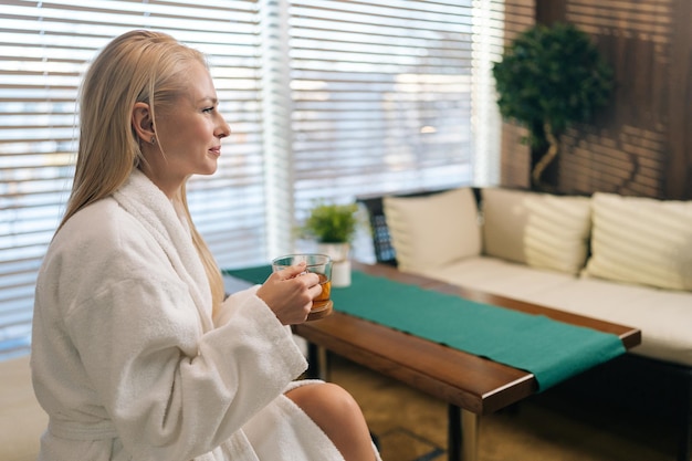 Vista lateral de una joven feliz y relajada con albornoz blanco disfrutando de té medicinal a base de hierbas frescas después de tratamientos de spa sentados en un sofá en el salón de spa