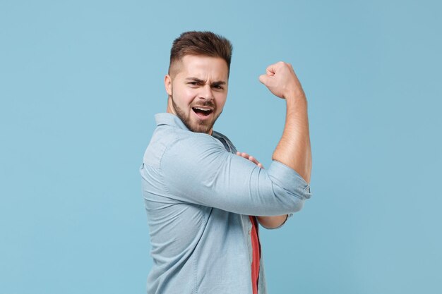 Vista lateral de un joven fuerte y barbudo de 20 años con un pantalones casual posando aislado en un retrato de estudio de fondo de pared azul pastel. Concepto de estilo de vida de las emociones de las personas. Simulacros de espacio de copia. Mostrando los músculos bíceps.