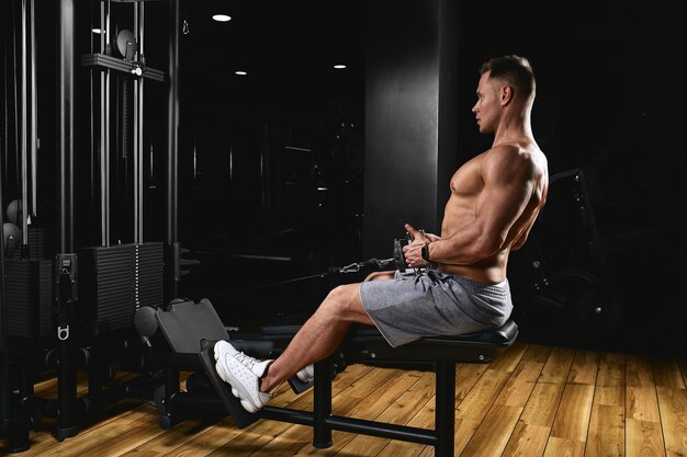 Vista lateral de un joven fuerte con cuerpo atlético haciendo ejercicios en aparatos de gimnasia en un gimnasio oscuro.