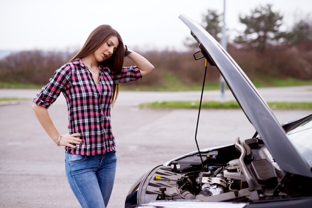 Foto vista lateral de una joven hermosa preocupada está mirando debajo del capó de su coche y sosteniendo su cabeza con desesperación.