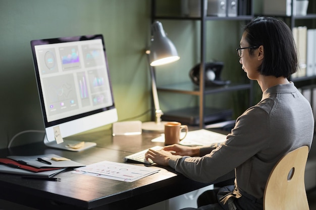 Vista lateral de un joven hombre de negocios asiático que usa una computadora con gráficos de datos mientras trabaja en la oficina de su casa