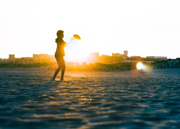 Foto vista lateral de un joven jugando al rugby en la playa contra un cielo despejado durante la puesta de sol