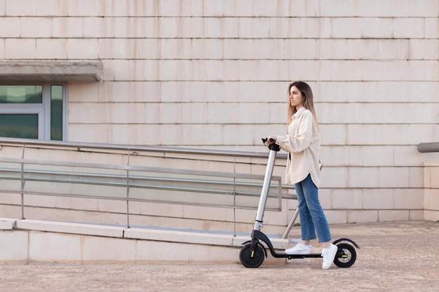 Vista lateral de una joven montando un scooter eléctrico sostenible en la ciudad frente a un edificio blanco con espacio para copiar