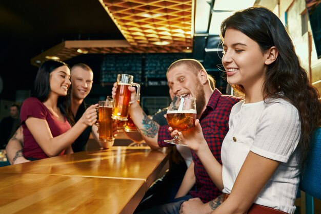 Vista lateral de la joven morena sonriente disfrutando de tiempo libre con amigos felices y bebiendo cerveza en el bar. Compañía alegre bebiendo alcohol, hablando y riendo en el pub. Concepto de bebida y diversión.