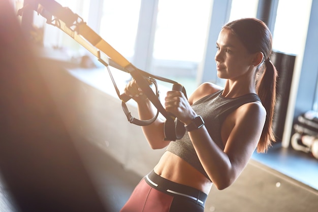 Vista lateral de una joven mujer fuerte y en forma haciendo ejercicio con correas de fitness trx en el gimnasio. Deporte, entrenamiento, bienestar y estilo de vida saludable