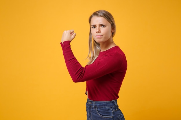 Vista lateral de una joven mujer rubia con ropa informal posando aislada en un retrato de estudio de fondo de pared naranja amarillo. Concepto de estilo de vida de las emociones de las personas. Simulacros de espacio de copia. Mostrando bíceps, músculos.