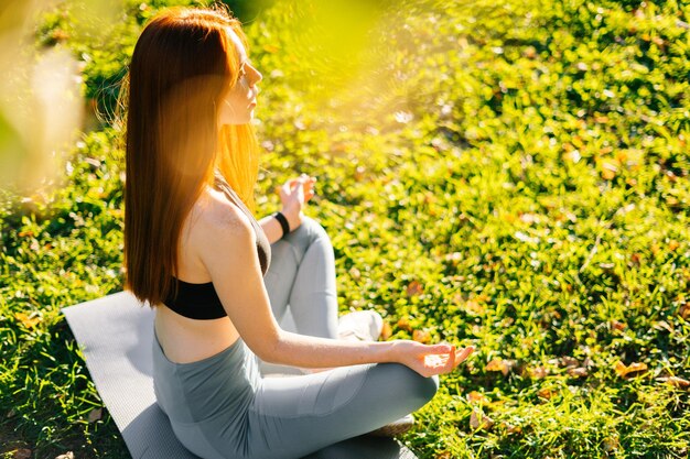 Vista lateral de una joven pelirroja con los ojos cerrados meditando en una pose de loto tomándose de la mano en la rodilla sentada en una alfombra de yoga sobre hierba verde en el parque de la ciudad en la soleada mañana de verano