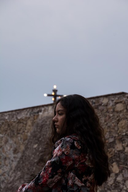Foto vista lateral de una joven pensativa que mira hacia otro lado mientras está de pie junto a la pared contra el cielo