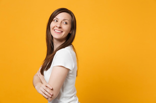 Vista lateral de una joven sonriente con ropa informal blanca con las manos cruzadas, mirando a un lado aislada en un fondo de pared naranja amarillo brillante en el estudio. Concepto de estilo de vida de las personas. Simulacros de espacio de copia.