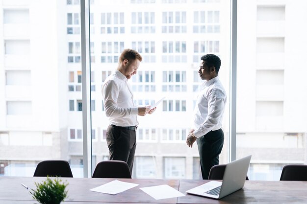 Vista lateral de jóvenes empresarios europeos afroamericanos y caucásicos discutiendo datos de documentos. Dos jóvenes empresarios multiétnicos exitosos en ropa formal.