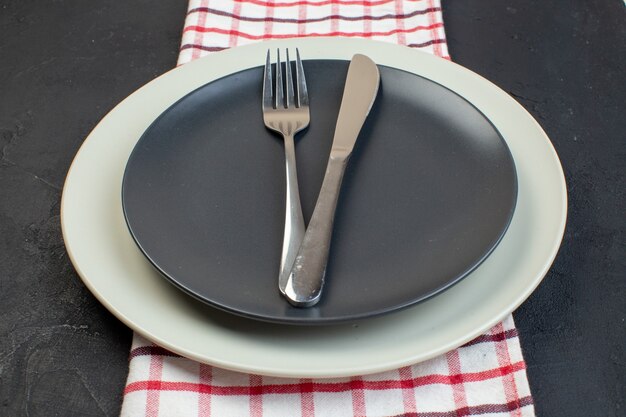 Vista lateral del juego de cubiertos de acero inoxidable en color gris oscuro y platos vacíos blancos sobre una toalla de rayas rojas sobre fondo negro con espacio libre