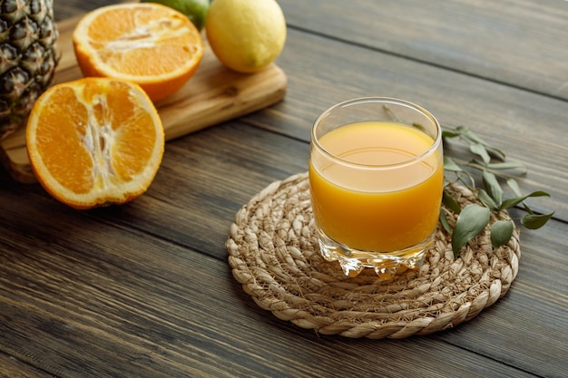 Vista lateral de jugo de naranja en vaso con hojas en salvamanteles y naranjas medio cortadas con piña de limón en tabla de cortar sobre fondo de madera