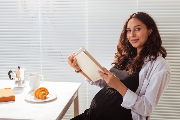 Vista lateral del libro de lectura feliz joven y bella mujer mientras desayuna con café y por la mañana