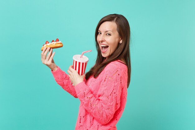 Vista lateral de la linda mujer joven en suéter rosa tejido sostenga en las manos pastel de eclair, vaso de plástico de cola o refresco aislado sobre fondo azul, retrato de estudio. Concepto de estilo de vida de personas. Simulacros de espacio de copia.