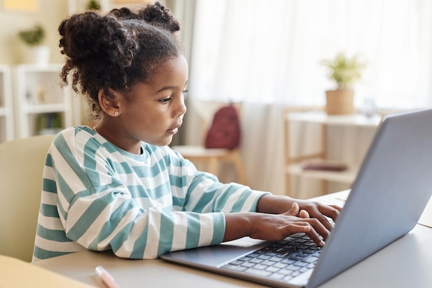 Vista lateral linda niña negra usando una computadora portátil estudiando en línea en casa