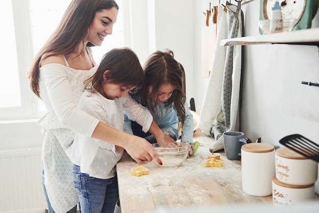 Vista lateral. Mãe e duas meninas na cozinha estão aprendendo a cozinhar boa comida com farinha