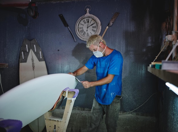 Vista lateral de un maestro adulto de cabello rubio con máscara respiratoria moliendo una tabla de surf blanca colocada en bastidores de conformación mientras trabaja en el taller