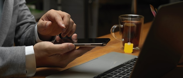 Vista lateral de las manos masculinas con smartphone mientras trabaja con la computadora portátil en la sala de la oficina