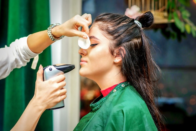 Vista lateral de un maquillador con aerógrafo haciendo una base de maquillaje con aerógrafo en un rostro femenino