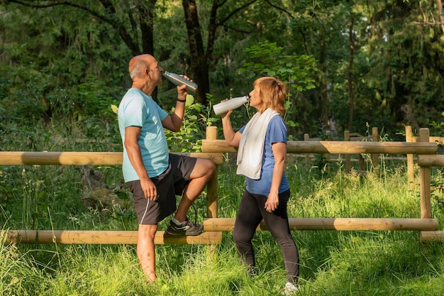 Vista lateral del marido y la mujer mayores bebiendo agua de una botella ecológica reutilizable en el parque