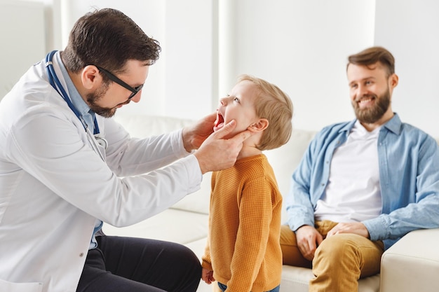 Vista lateral del médico masculino que inspecciona la garganta del niño pequeño cerca del padre durante la cita de control en el hospital contemporáneo