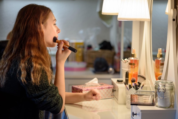 Foto vista lateral de una mujer aplicando maquillaje frente al espejo en casa
