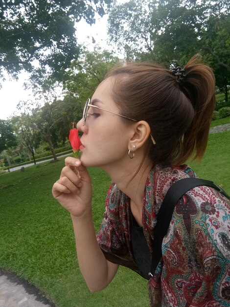 Foto vista lateral de una mujer besando una flor roja mientras está de pie en el parque