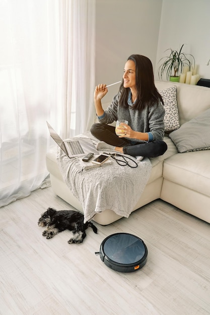 Vista lateral de una mujer con cabello castaño sentada con un vaso de bebida en un sofá con una computadora portátil abierta y un teléfono inteligente en una habitación con un perro dormido y una aspiradora robótica