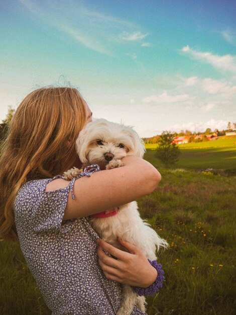 Foto vista lateral de una mujer cargando un perro mientras está de pie en tierra contra el cielo