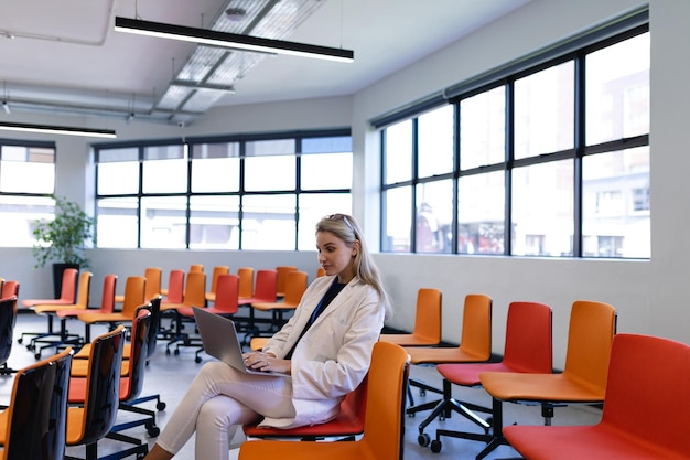 Vista lateral de una mujer caucásica con cabello largo y rubio, vestida con ropa elegante, sentada en una silla en una moderna sala de reuniones vacía, usando una computadora portátil.