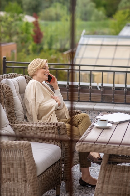 Vista lateral de una mujer elegante rubia contenta sentada con las piernas cruzadas en el sillón en una mesa de madera en la terraza