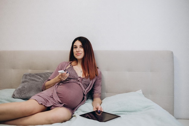 Vista lateral de una mujer embarazada relajada viendo películas en la televisión Una mujer morena con ropa doméstica se sienta en un sofá con un plato con palomitas de maíz