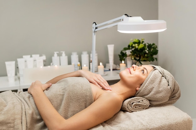 Vista lateral de una mujer encantada con toallas en la cabeza y el cuerpo acostada en la cama bajo una lámpara en un centro de belleza