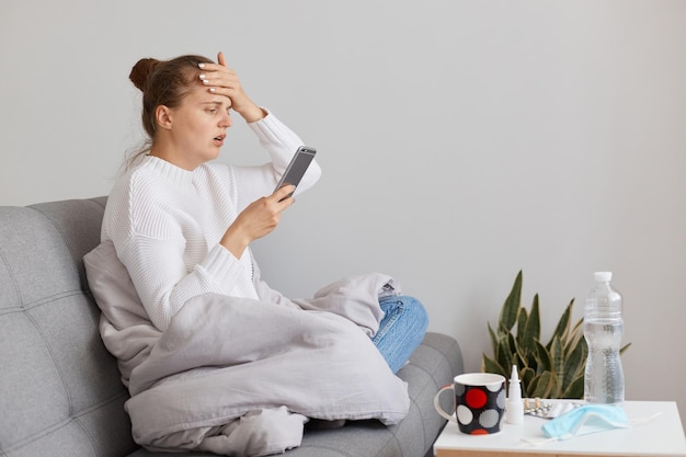 Vista lateral de una mujer enferma que usa suéter blanco y está envuelta en una manta tocándose la frente con dolor de cabeza y alta temperatura sosteniendo un teléfono inteligente en las manos