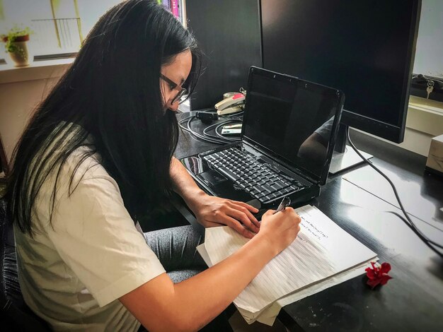 Vista lateral de una mujer escribiendo código informático junto a una computadora portátil y un monitor en la mesa