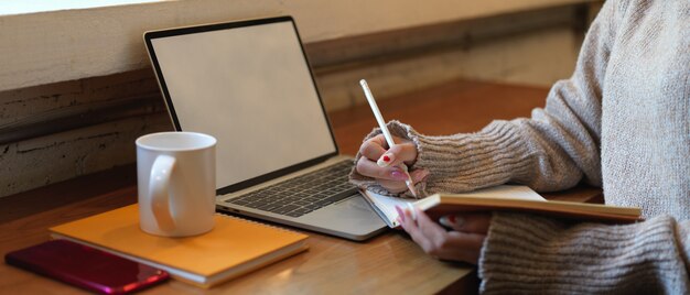 Vista lateral de la mujer escribiendo sin cuaderno en blanco mientras se encuentra en la mesa de trabajo de madera con una computadora portátil y un teléfono inteligente simulados