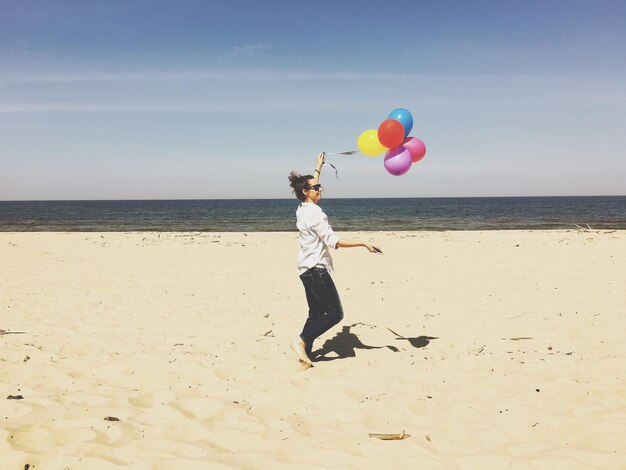 Vista lateral de una mujer feliz sosteniendo globos coloridos mientras camina por la arena en la playa durante un día soleado